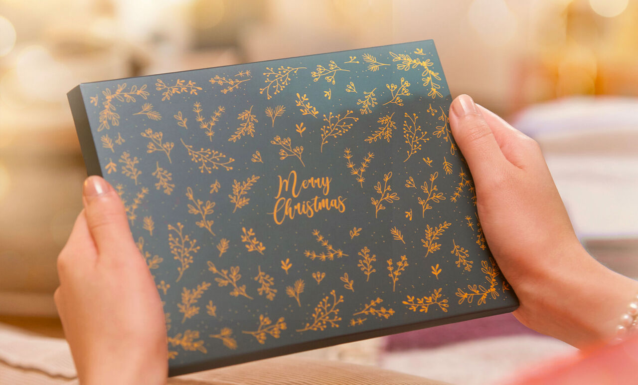 Manos sosteniendo una caja de regalo oscura con motivos florales dorados y el texto Merry Christmas.