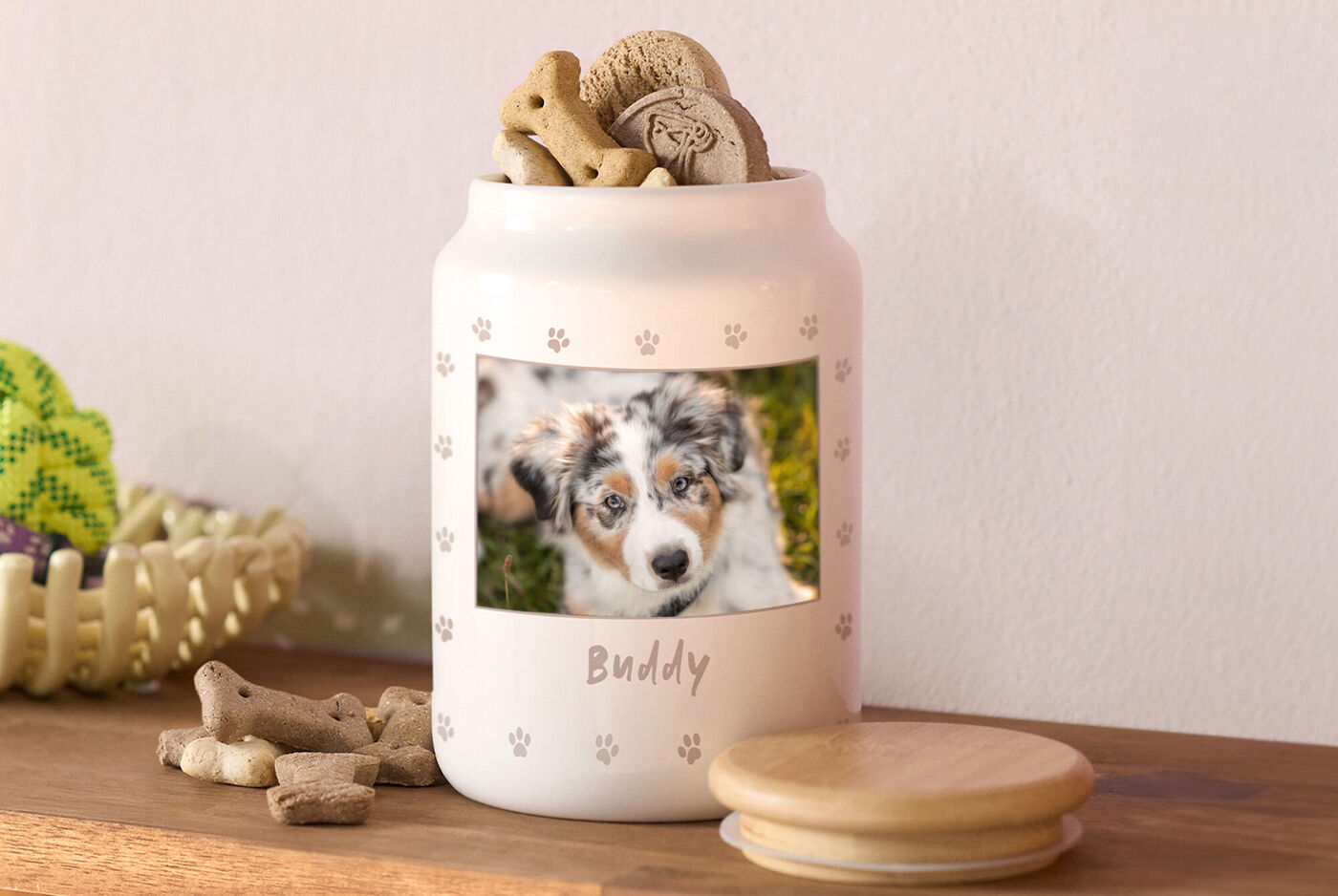 Tarro de cerámica personalizado con foto de perro y nombre "Buddy", rodeado de snacks para mascotas sobre una superficie de madera.